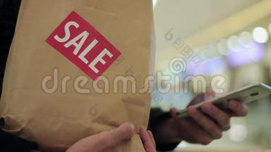男士一只手拿着带有销售标签的棕色纸袋，在商场里使用智能手机